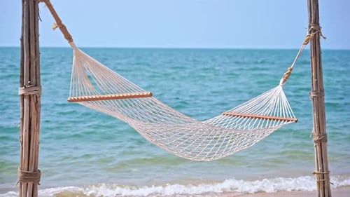 Empty white hammock hangs from poles on tropical island beach with calm turquoise sea in background