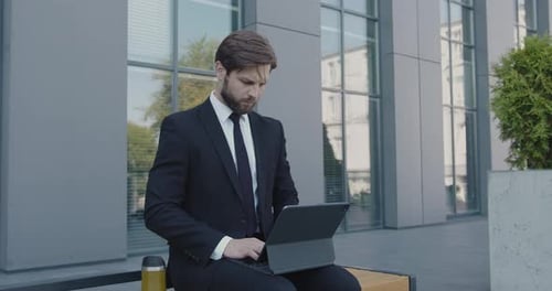 Ernsthafter bärtiger Geschäftsmann arbeitet auf Bank mit Laptop im Freien