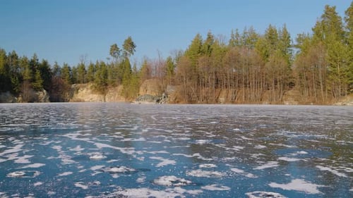A frozen lake with a textured icy surface reflects the clear blue sky. Surrounding it tall evergreen