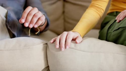 Midsection of diverse couple sitting on sofa holding hands at home, in slow motion