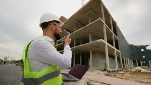 Male Architect Using Laptop and Walkie Talkie at Construction Site