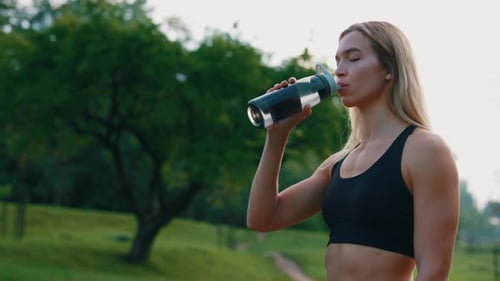 Side View of the Caucasian Female Runner Standing in the Park Drinking Water From the Sport Bottle