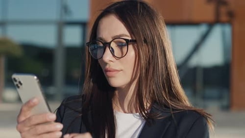 A Beautiful Young Businesswoman with Glasses Using a Modern Smartphone Stand in the City Looking at