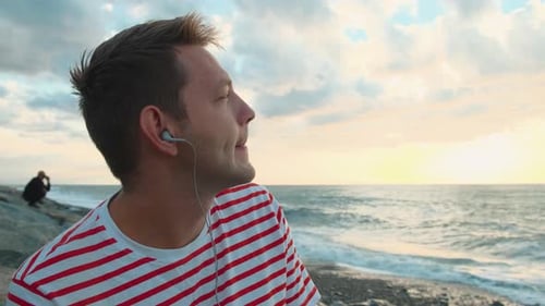Happy Caucasian Young Man Listening to Musc with Headphones Using Smartphone on the Sea Coast and