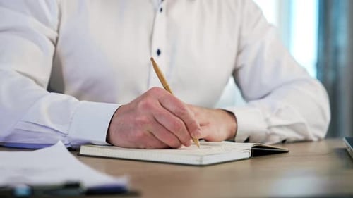 Manos de un hombre vestido con camisa blanca tomando notas en el libro de papel.