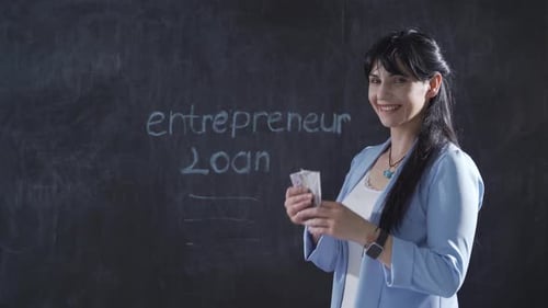 Woman writing entrepreneur loan on blackboard looks up with happy expression.