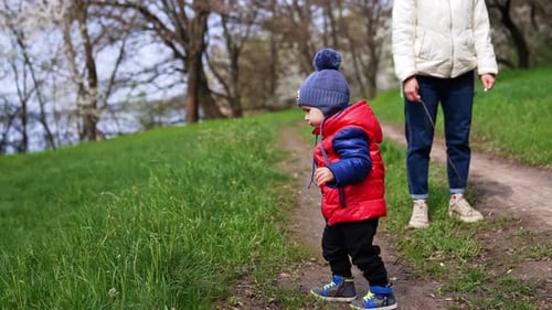 Little toddler on the walk in the nature with his mom. Baby boy in red blazer goes to green grass.
