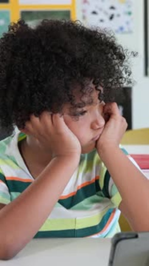 Vertical video: Resting kid in striped tee gazing at tablet on table, papers, red folder studying