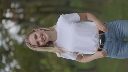 Vertikale Aufnahme einer selbstbewussten und glücklichen blonden Frau, die sich in einem Park entspannt und direkt in die Kamera schaut