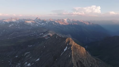 Dramatic drone aerial of snow-flecked alpine ridge at sunset, descending into a deep mountain valley