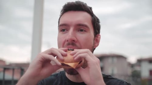 Slow motion young caucasian man bites into burger at home, cheeseburger eating enjoyment, close up p