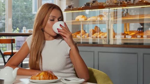 Woman Drinks Coffee and Eats Croissant in Cafe