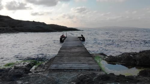 Die Aufnahme bewegt sich bei Sonnenuntergang nach unten und horizontal entlang eines Holzpfiers von der Küste zum Meer.