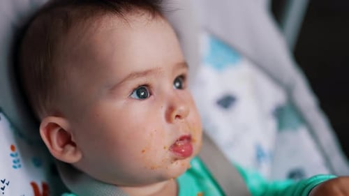 Caucasian baby dirtied with food after feeding. Little kid shoves the spoon into mouth. Close up.