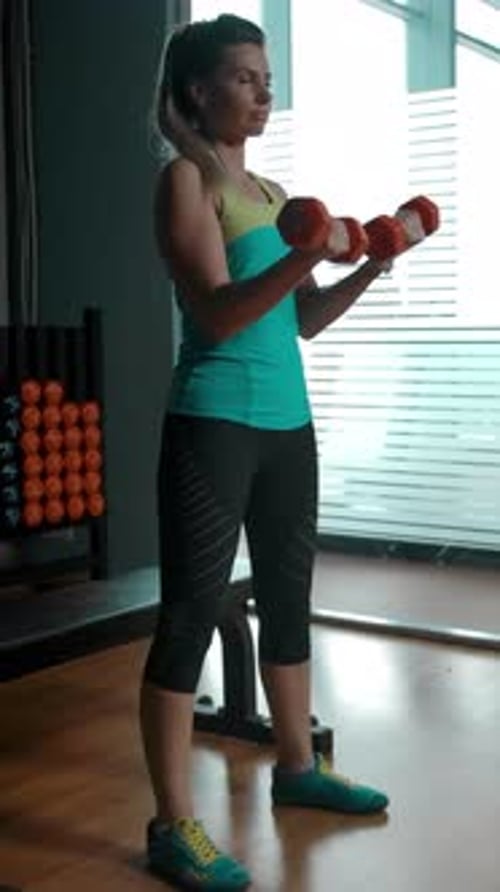 Vertical of Caucasian Blonde Woman Doing Exercises for Biceps with Two Dumbbells in the Gym