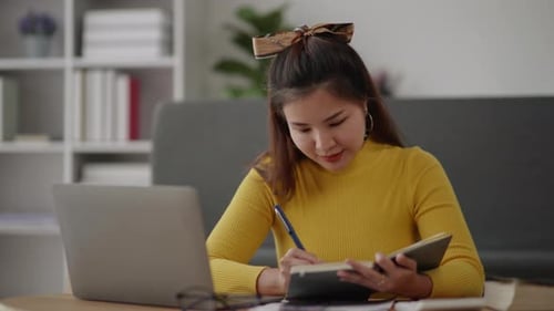 Woman Writing in Notebook Next to Laptop at Home