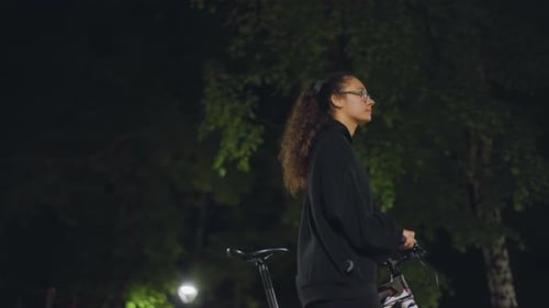 Young Lady With Bike Halts Beneath Nighttime Park Illumination Silhouetted Young Woman With Bicycle