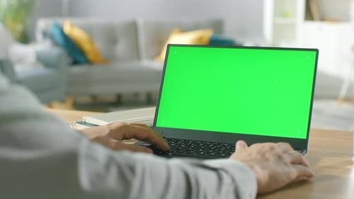 Close-up of a Man Uses Laptop with Green Mock-up Screen While Sitting at the Desk in His Cozy Livin