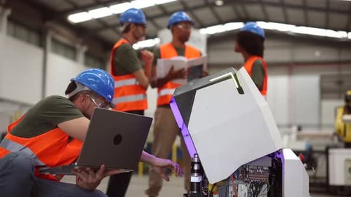 Multiracial team of engineers programming a robot using a laptop in a modern industrial factory, col