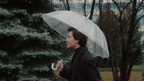 Young Man in a Coat Unhurriedly Walking Under a Transparent Umbrella in the Autumn City Park Looking
