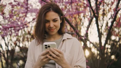 Attractive Young Woman Uses Smatphone in Park at Sunset Spring Flowers of Cherry or Sakura Blossoms