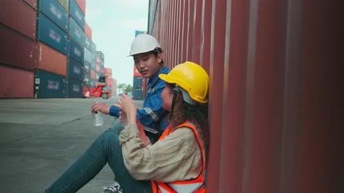 Two Asian workers rest from work drink water and relax at stacks of containers.