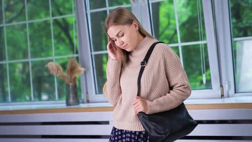 Young blonde woman in beige sweater standing indoors at the window.