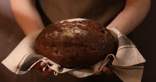 Woman holding loaf of fresh rye bread on brown background, closeup