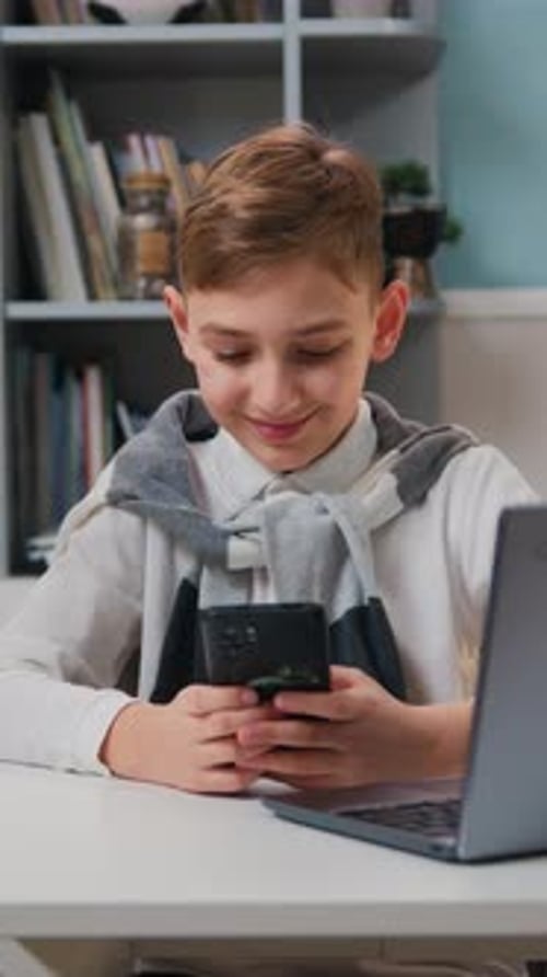Smiling Boy Using Smartphone at Desk with Laptop