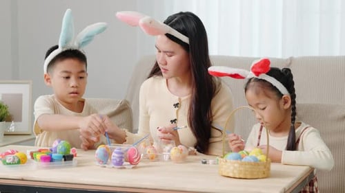 A Mother and Kids with Bunny Ears Happily Paint Easter Eggs at a Table Near a Festive Basket