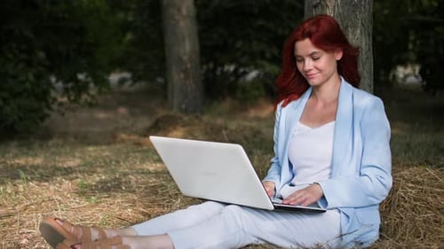 Remote Work a Young Businesswoman Works at Computer in a Park Sitting in a Clearing Among Trees