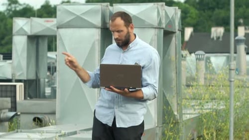 Young technician with laptop analyzing air conditioning system on city roof
