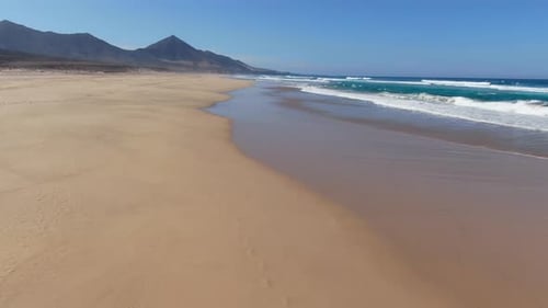 Low altitude drone flight over a deserted sandy paradise beach with blue waves. Beautiful mountains