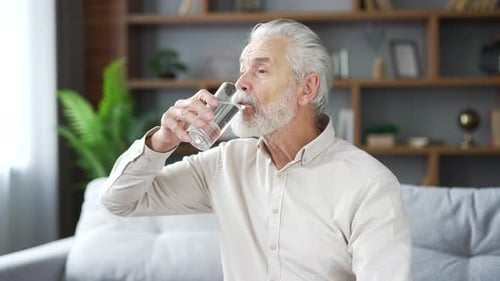 Älterer älterer grauhaariger bärtiger Mann trinkt Wasser aus einem Glas und sitzt auf dem Sofa im Wohnzimmer