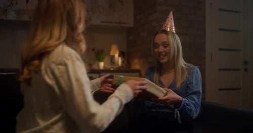 Happy Blonde Birthday Girl in a Festive Cap Receives a Gift with a Bow From Her Girlfriend During a