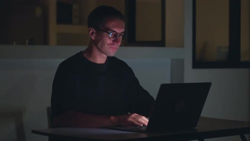 Mann arbeitet spät in der Nacht am Laptop und tippt Code mit fokussiertem Ausdruck und Nachtarbeit bei schlechten Lichtverhältnissen