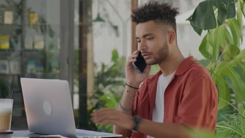 Man Using Laptop and Talking on Phone in Street Cafe