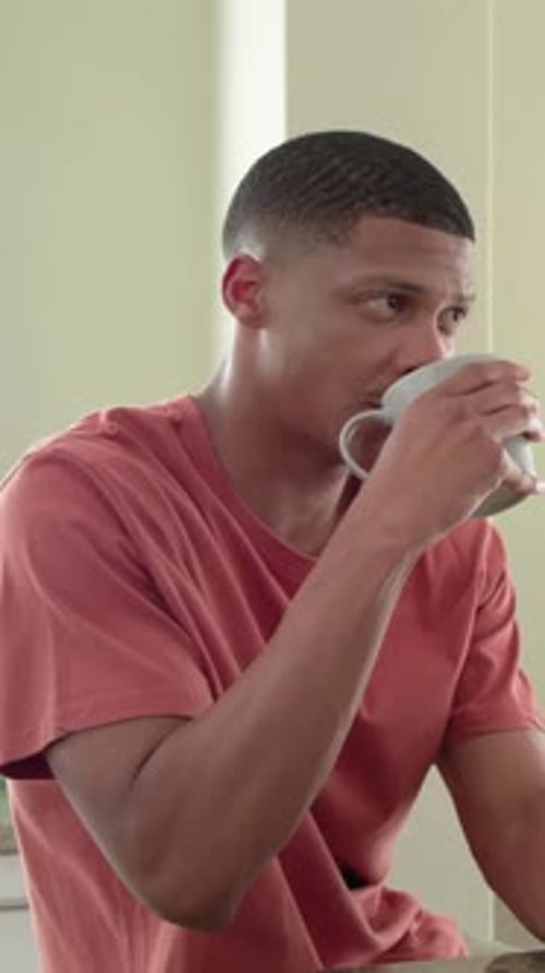 Vertical video: Hearing voice, he in coral tee with band reacting sipping mug at counter, smiling