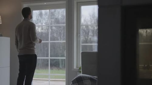 Casual man drink coffee and looking to cold outside from cozy home, back view