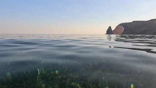 Sea Water Surface Camera Flies Over the Calm Azure Sea with Volcanic Rocky Shores on Background