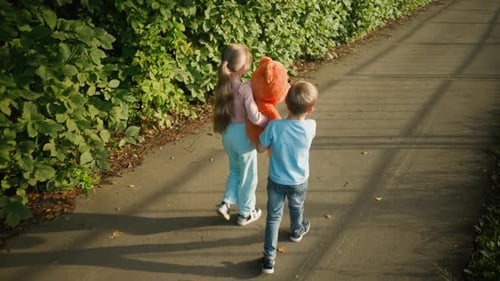 Boy and Girl Walking on Path Adjusting Teddy Bear with Shadows on Ground