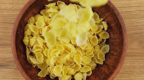 Shell-shaped pasta is poured into a brown ceramic bowl against a wooden oak board. Top view. Slow