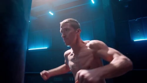 Young Male Kickboxer Punching Fists and Kicking Legs Punching Bag in Dark Gym with Blue Light