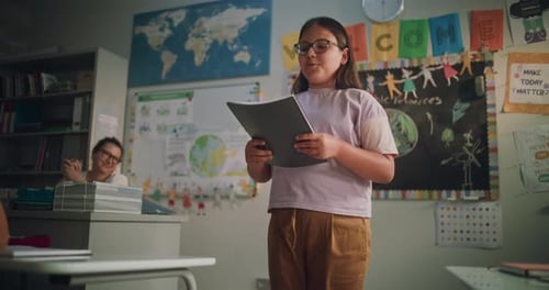 Primary School Girl Holding Notebook Showcasing Knowledge of Geography in Front of Classmates