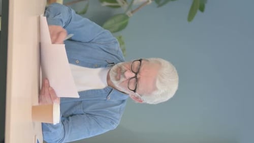 Senior Man Reviews Documents at Desk, Vertical Format
