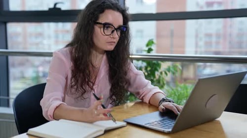Businesswoman sitting at table in a spacious office, working with laptop and taking notes.