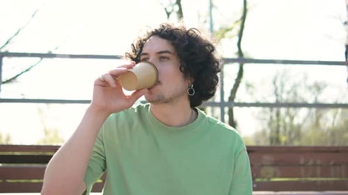 Close Up Shot of Young Curly Brunette Man Sitting in Park Drinking Morning Coffee