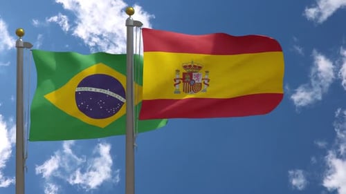 Waving Brazil And Spain Flags On Flagpoles Against Blue Sky