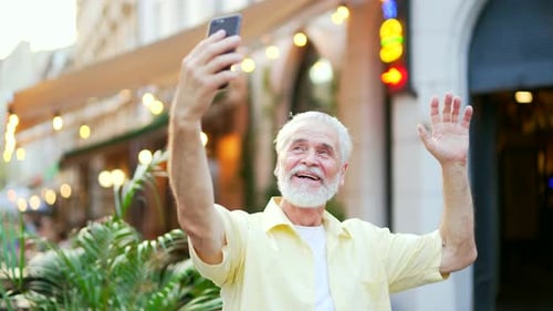 Senior gray haired bearded man talking on a video call using a smartphone while standing on a city