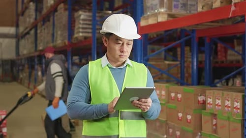 Asian Warehouse Manager in Helmet Texting on Digital Tablet